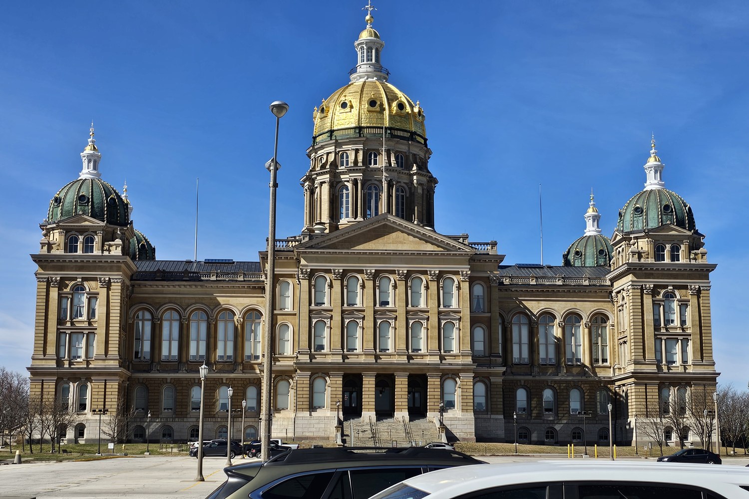 The Iowa State Capitol in Des Moines on a clear day: a tan stone building with a large gold central dome and four smaller green domes, with parked cars in the foreground.